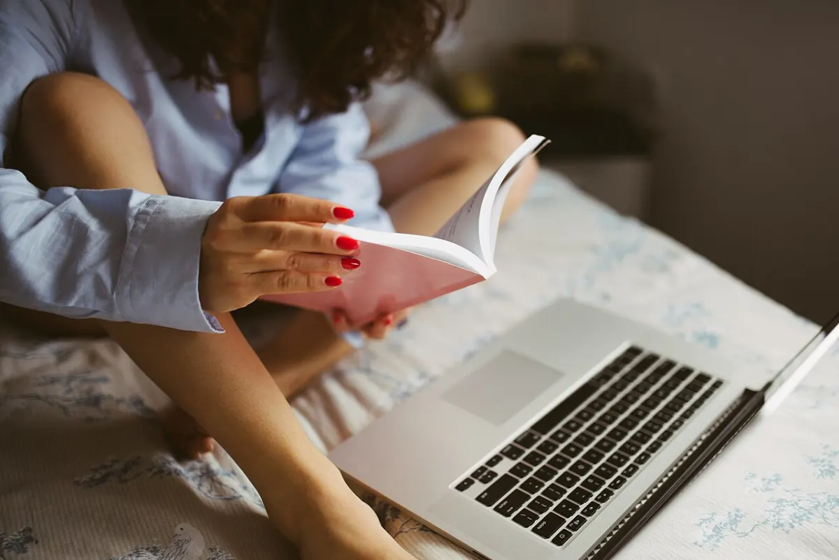 Book Review Website - The girl is reading a book on the bed and working on laptop