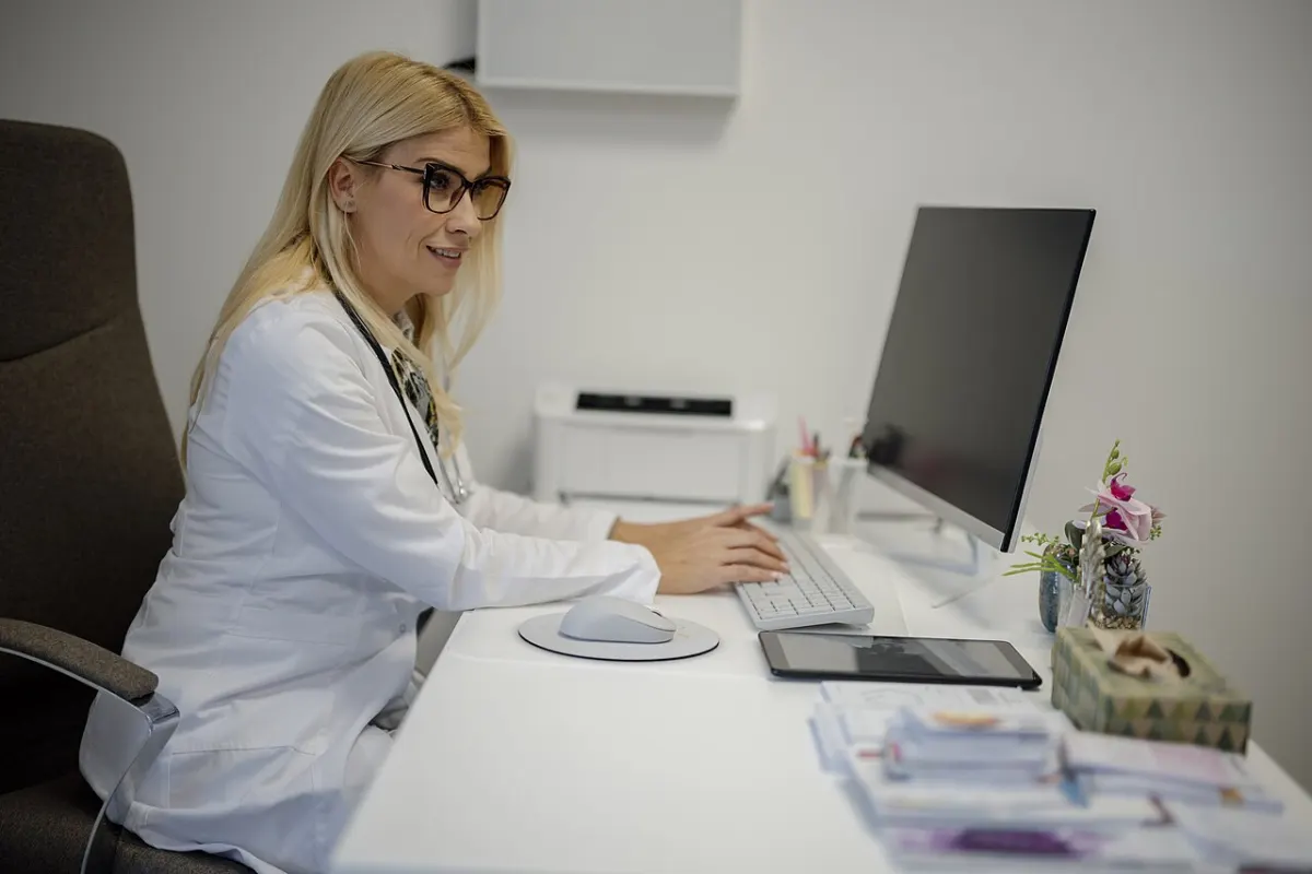 Purchase Order Management System - Doctor working at a desk in a medical office setting with a computer and stationery